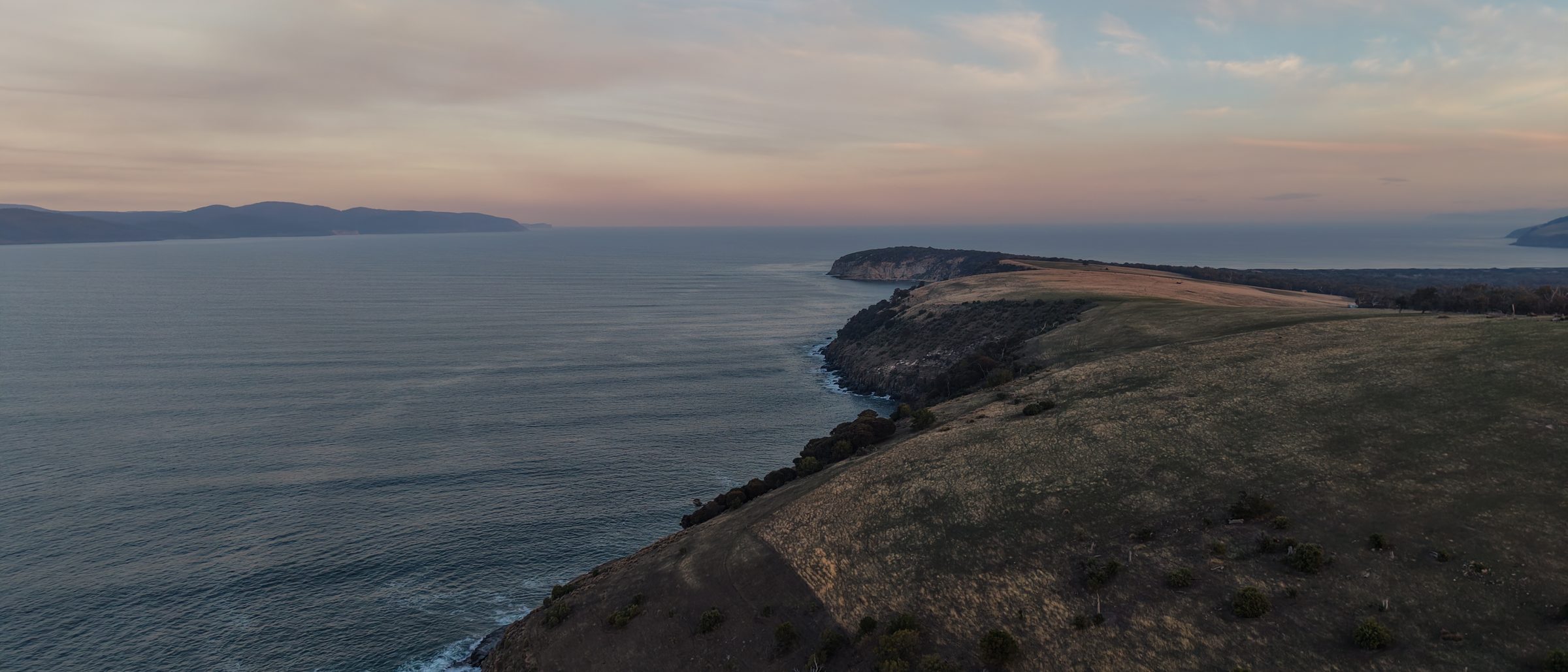 A Tasmanian headland at dusk, seen from the air: grass hills falling to a calm sea under a pink and blue sky.